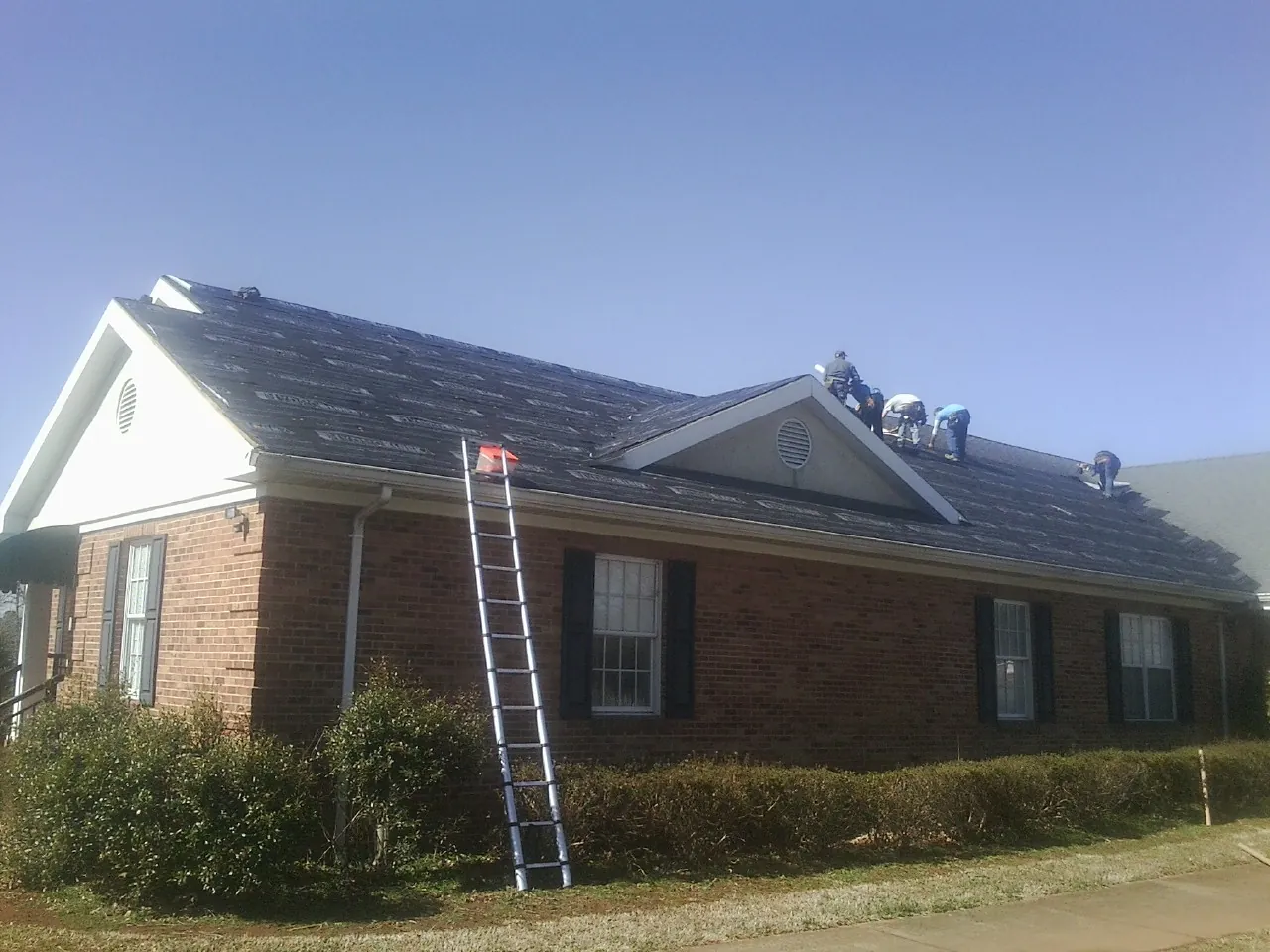 Crew tearing shingles off a brick ranch roof with a ladder set — Metal Roof Repair work by our crew in Henry